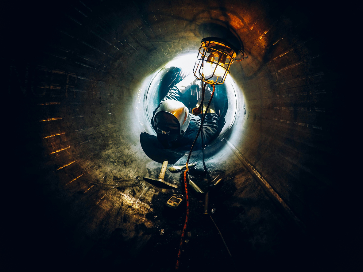 Worker in safety gear crawling through confined space tunnel with light