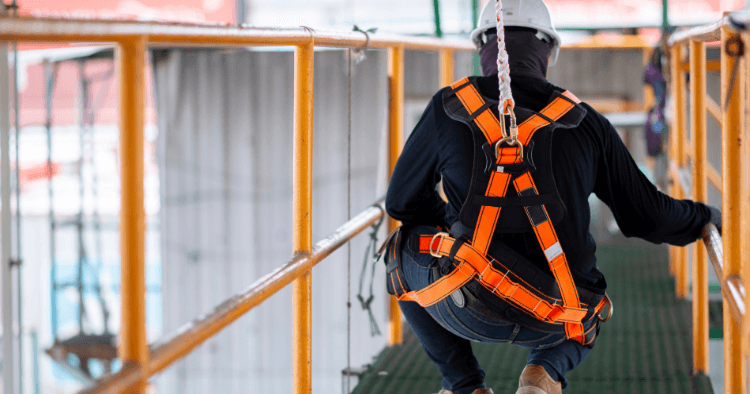 Worker in an orange fall arrest harness clipped to an anchor point while descending a set of stairs on a job site.
