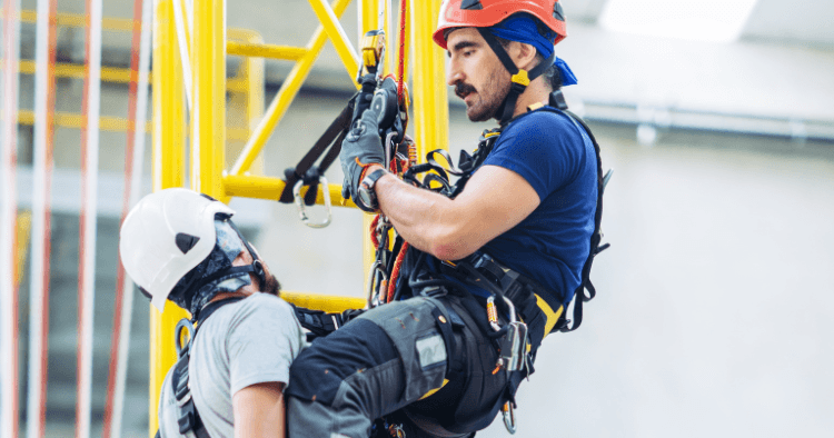 Two rope rescue technicians in helmets performing a suspended rescue technique, with one worker supporting the other during descent.
