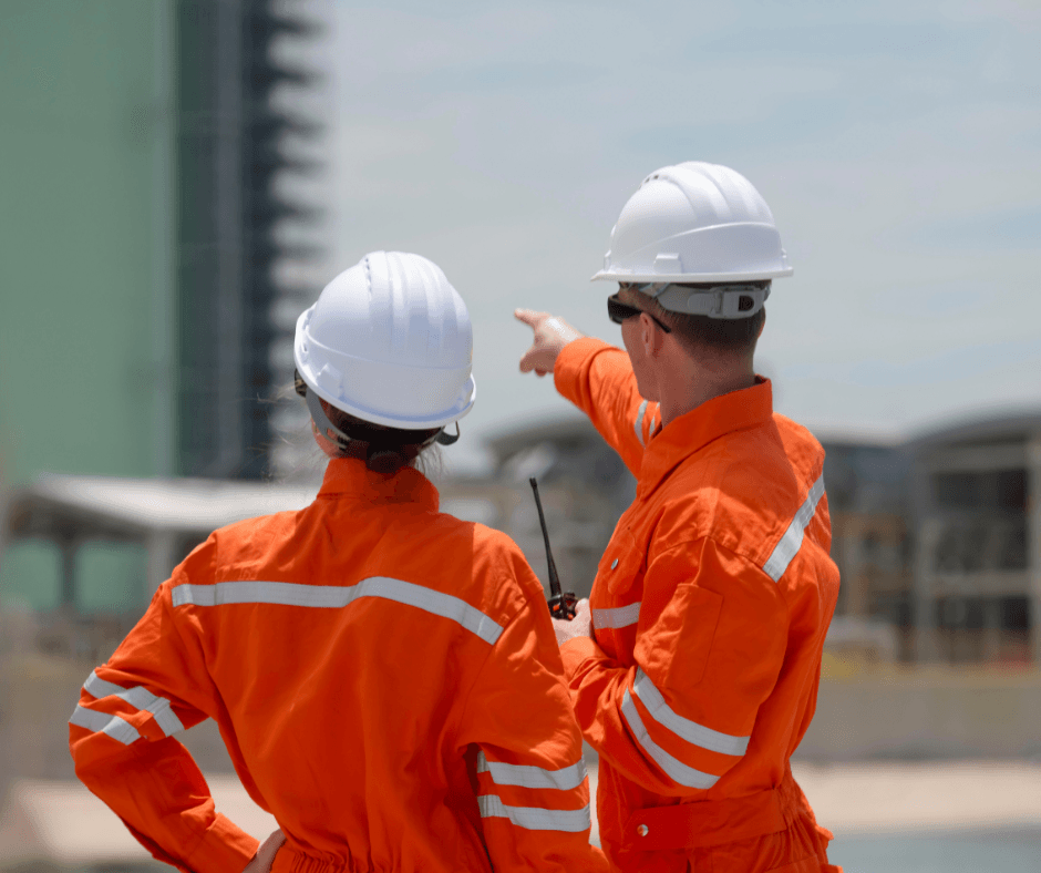 Two workers in orange high-visibility coveralls and white hard hats stand with their backs to the camera at an industrial site, one pointing toward a structure in the distance.
