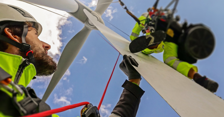 Two rope access technicians in helmets and harnesses work on a wind turbine blade, one suspended on lines beside it