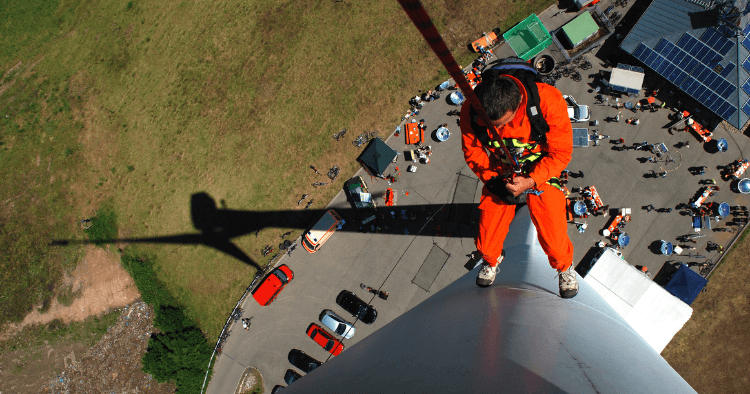 Turbine technician in an orange suit stands atop a tower, secured by rope, high above vehicles and crew on the ground