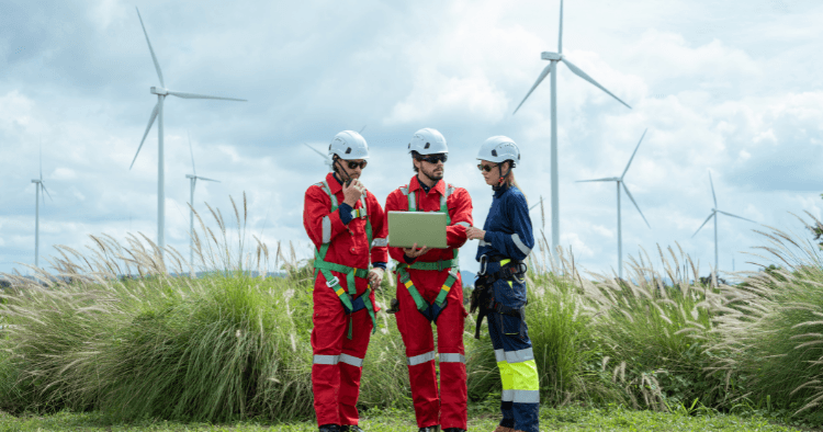 Three wind technicians in helmets and fall protection harnesses review a laptop in a field of wind turbines