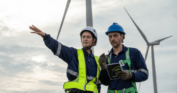 Two turbine workers in hi-vis vests and harnesses review a tablet while one points toward nearby wind turbines