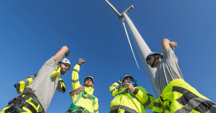 Four climbers in hi-vis jackets and helmets raise their arms beneath a wind turbine tower under a clear blue sky
