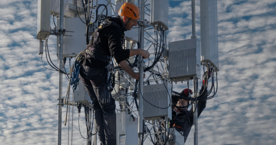 Two workers in fall-protection harnesses perform maintenance on cell tower equipment, one wearing an orange safety helmet while accessing antenna wiring against a cloudy sky.