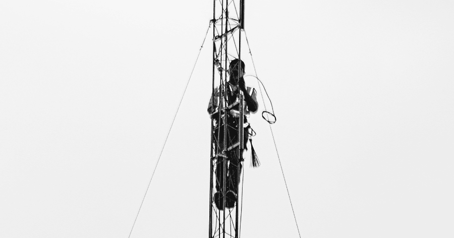 Black and white photo of a tower technician in a full-body harness climbing a lattice tower, guy wires visible against a plain sky background.