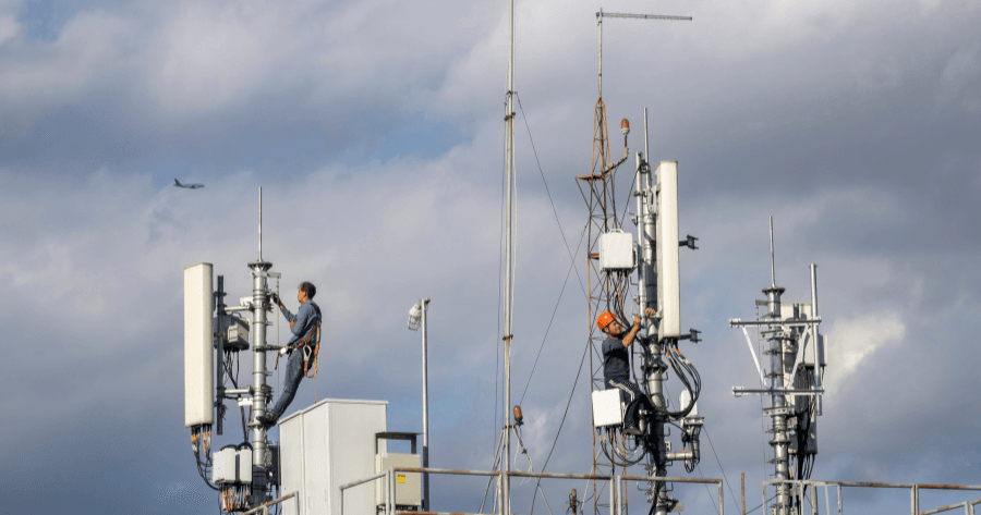 Two technicians work at height on a rooftop small cell installation, one perched on equipment near an antenna mount while a plane passes in the cloudy sky behind them.