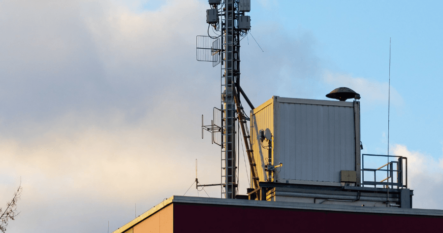 A rooftop small cell site with an antenna mast, dish, and HVAC unit crowded together against a partly cloudy sky.