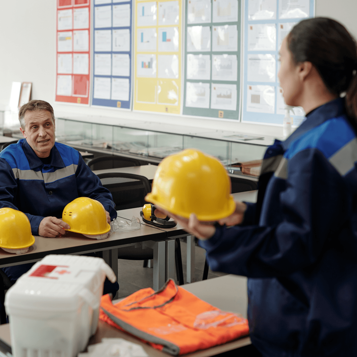 A safety instructor in blue workwear reviews hard hats and PPE equipment with a worker during an indoor training session.