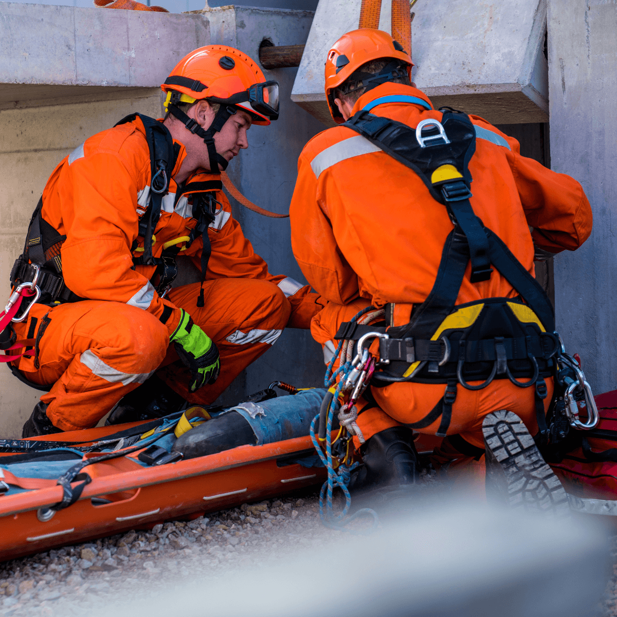Two rescue workers in full orange PPE and harnesses attend to a person on a stretcher during a technical rescue scenario.
