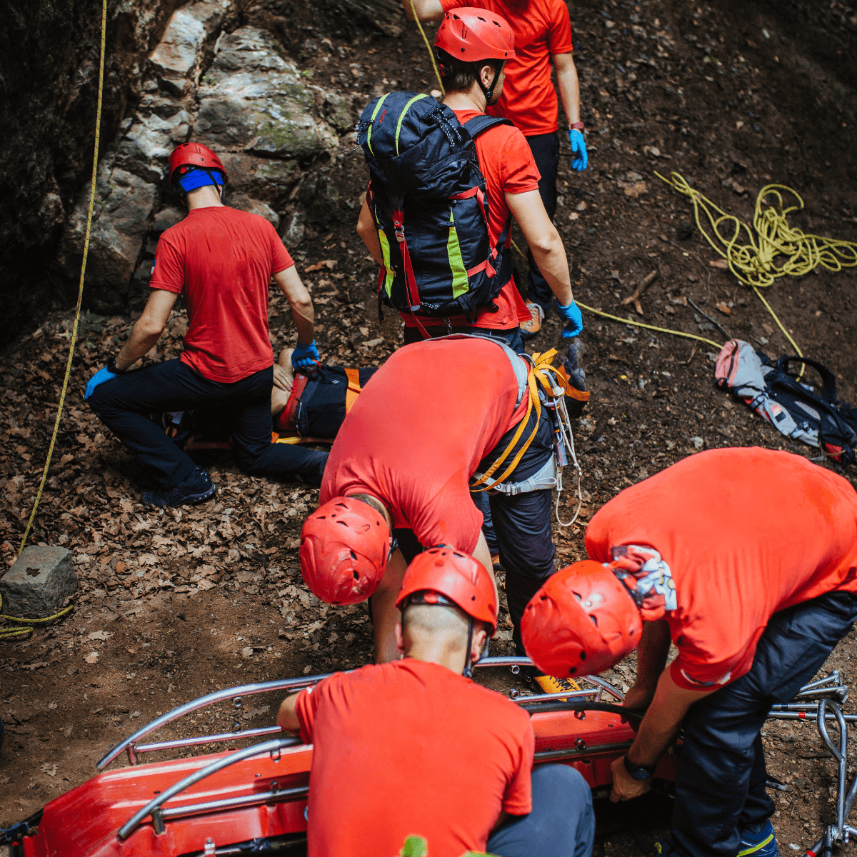 A team of rescue workers in red helmets and harnesses work together to move a patient on a stretcher through rough terrain.