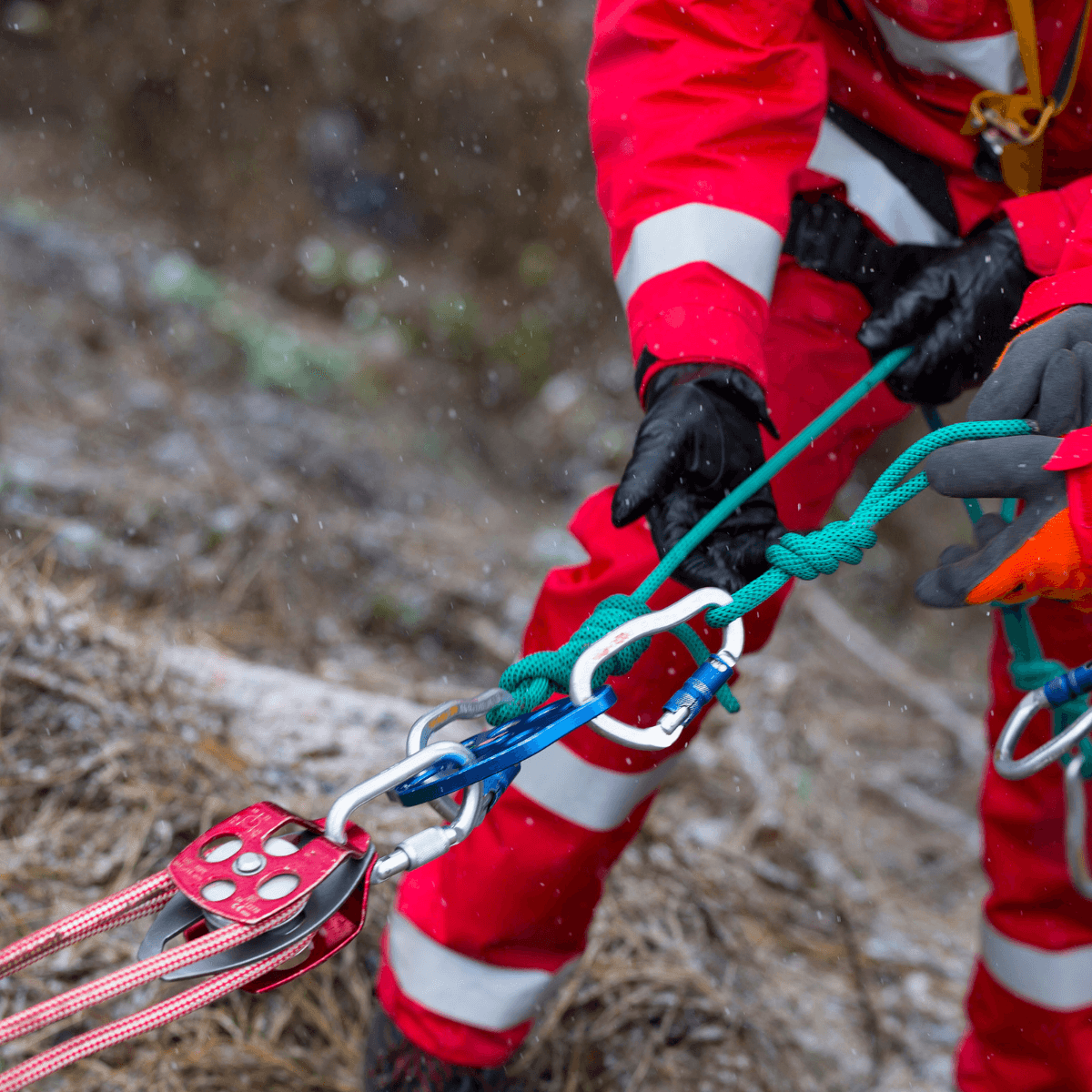 A rescue worker in a red high-visibility suit and black gloves manages a rope rescue system with carabiners and a pulley on a rocky outdoor terrain in wet conditions.