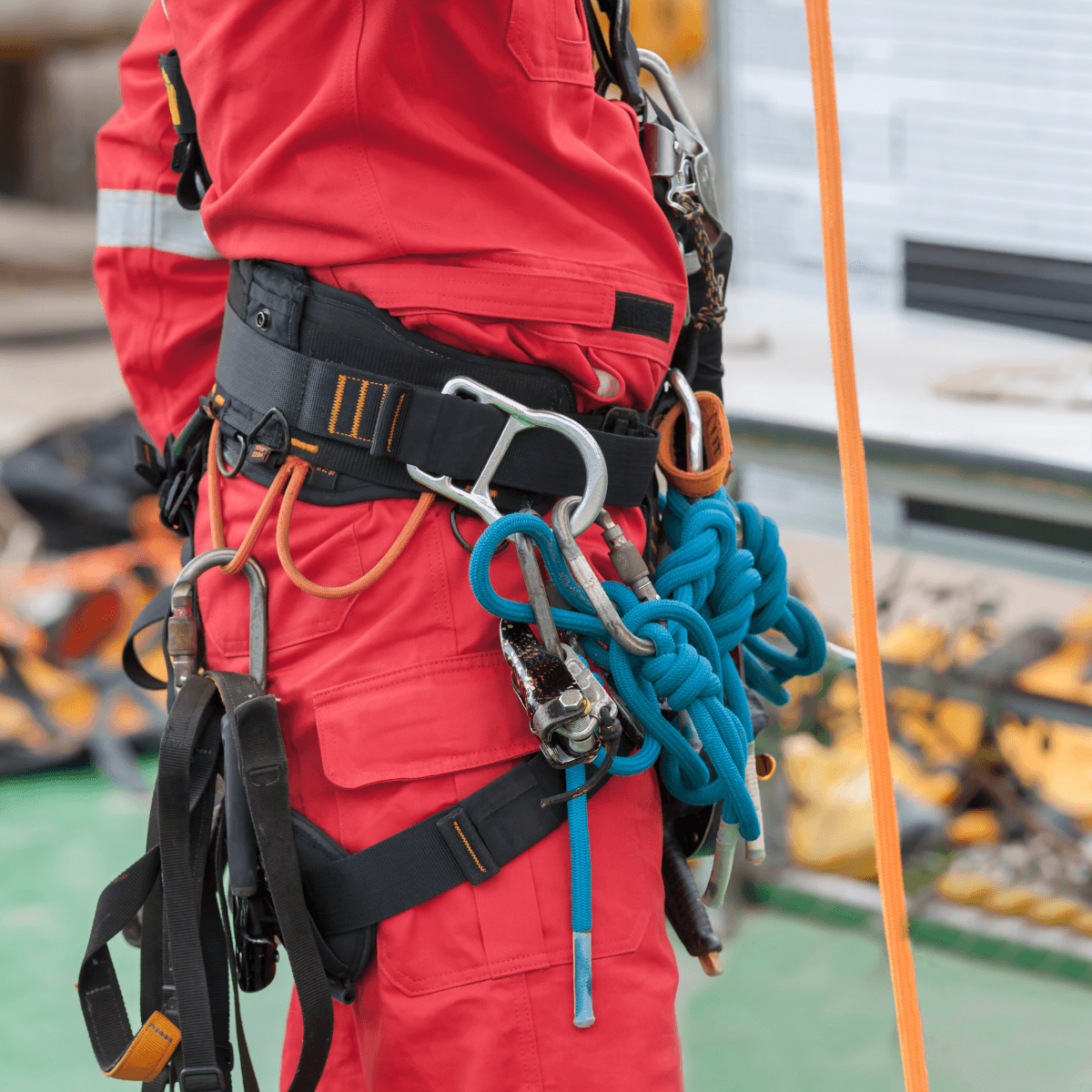 A close-up of a rescue worker's torso in red coveralls, showing a fully loaded harness with multiple carabiners, coiled ropes, and rigging equipment clipped to a safety belt.
