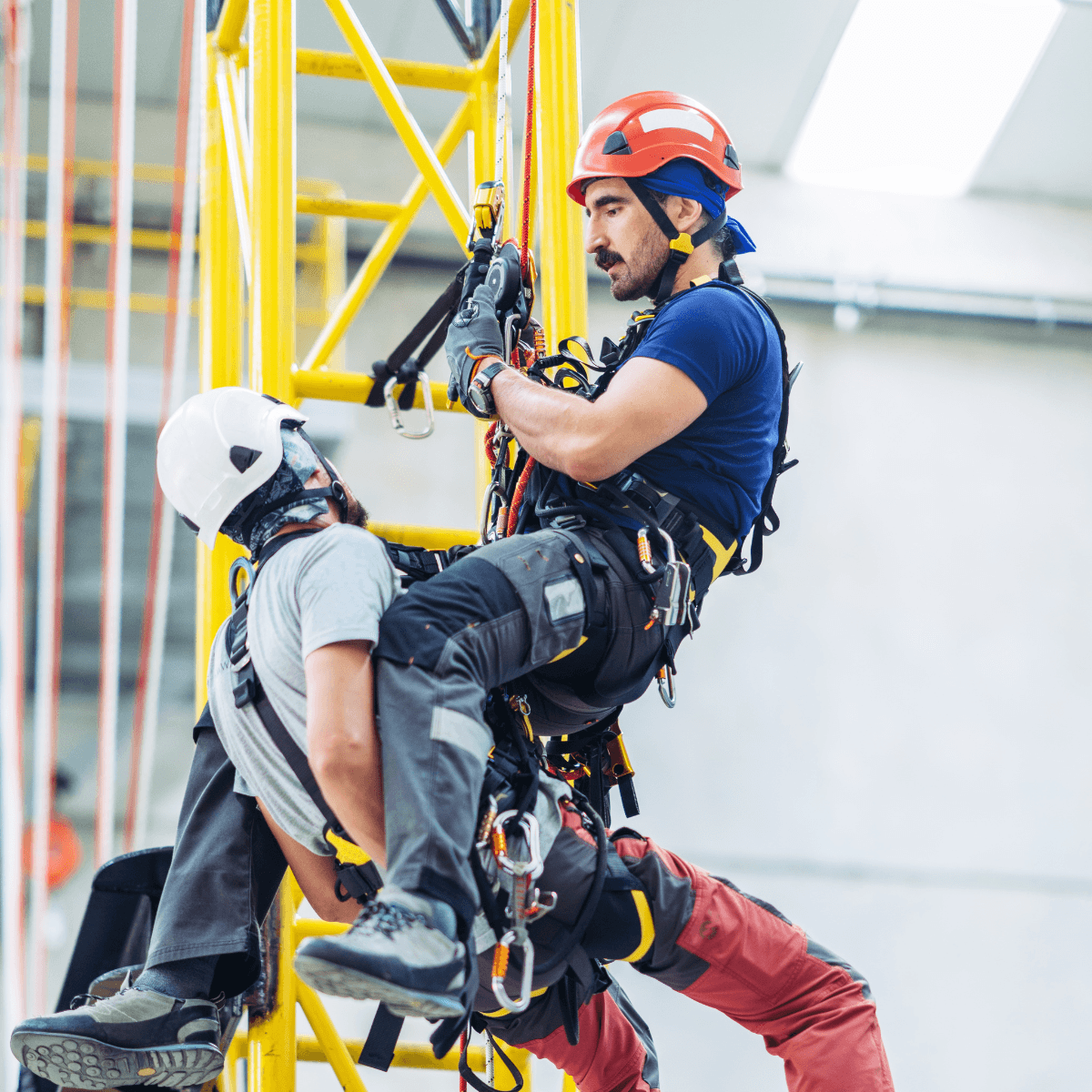 A rope rescue technician in a red helmet and full harness supports an incapacitated worker in a white helmet while suspended from a yellow industrial structure during a high-angle rescue training exercise.