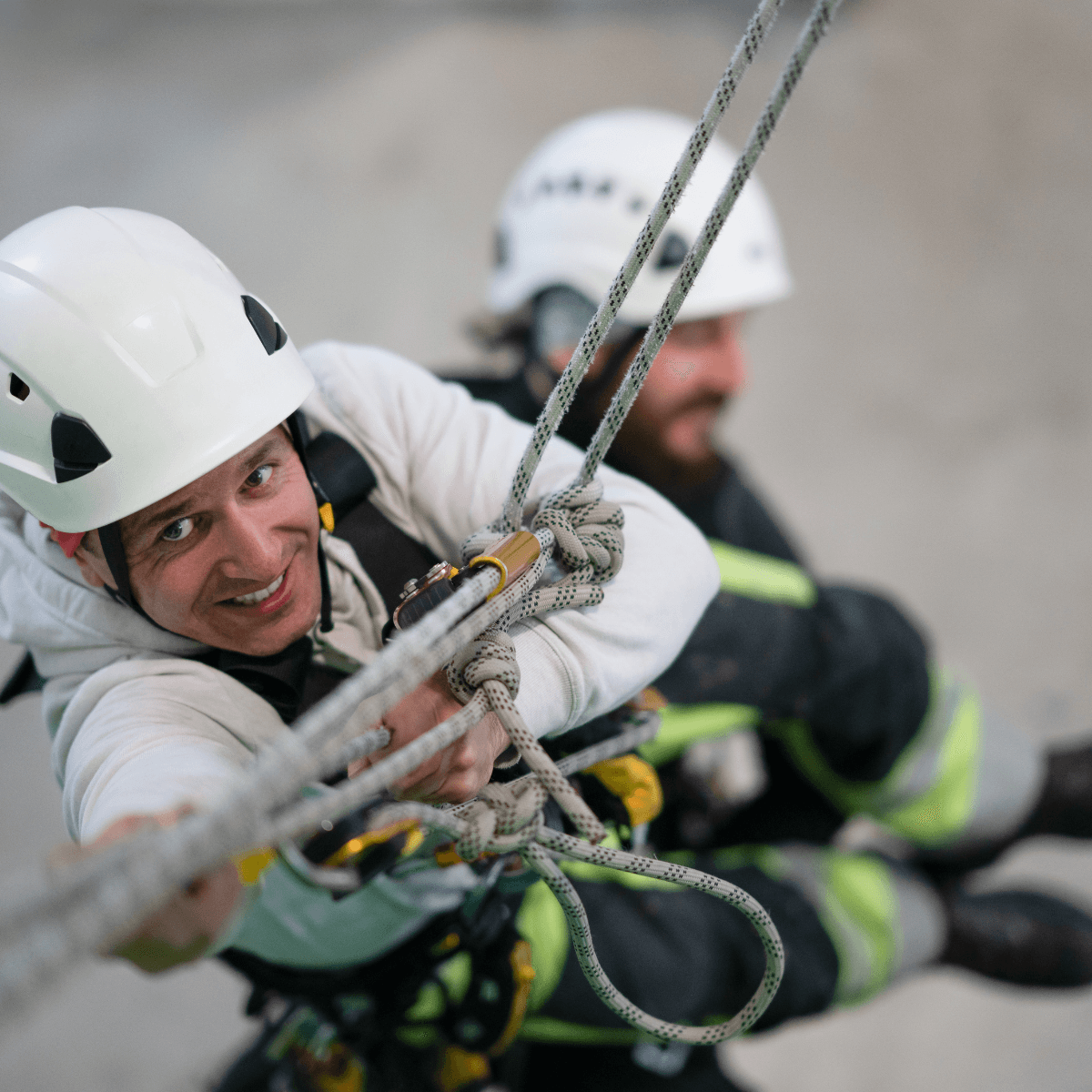 A rescue worker in a white helmet grips a rope system with focused intensity during a rope rescue drill, with a second helmeted responder visible in the background.