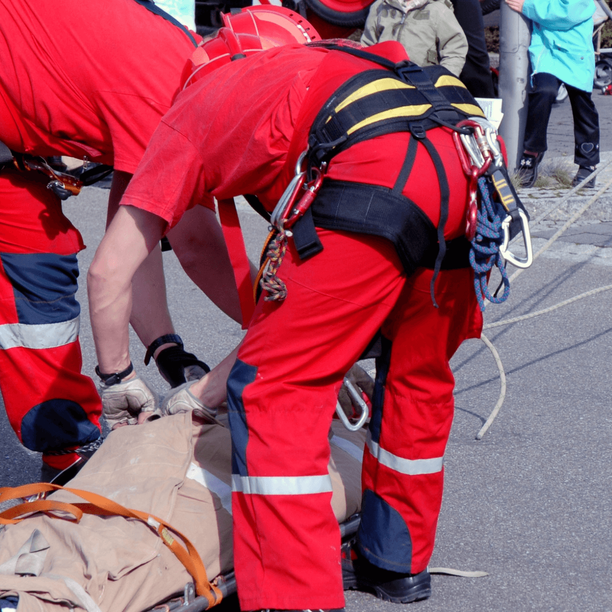 Two rescue workers in red uniforms and rope rescue harnesses lean over a person on a stretcher on an outdoor paved surface, securing the patient during a rescue demonstration.