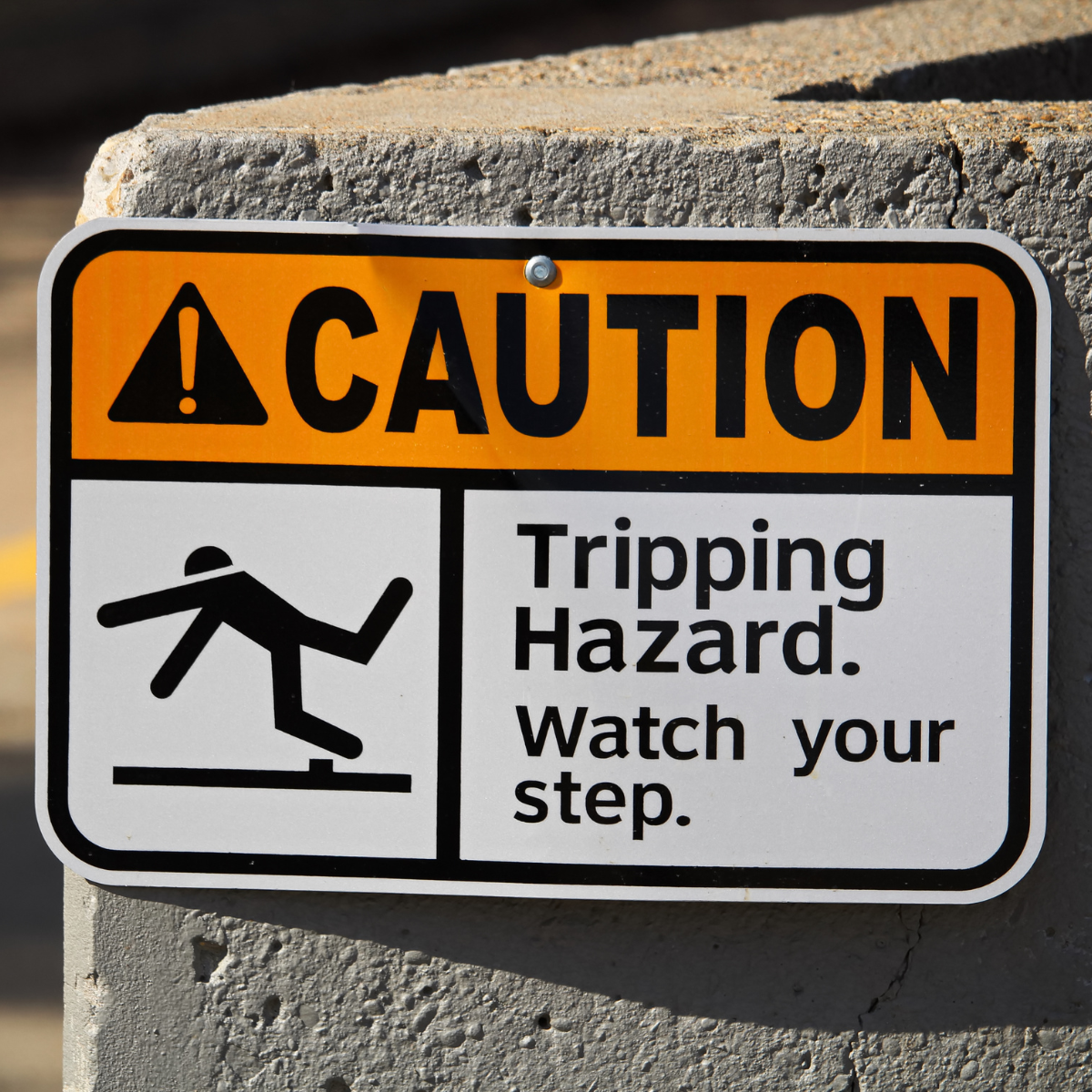 A orange and white caution sign mounted on a concrete pillar, warning of a tripping hazard with a stick figure stumbling over a raised surface.