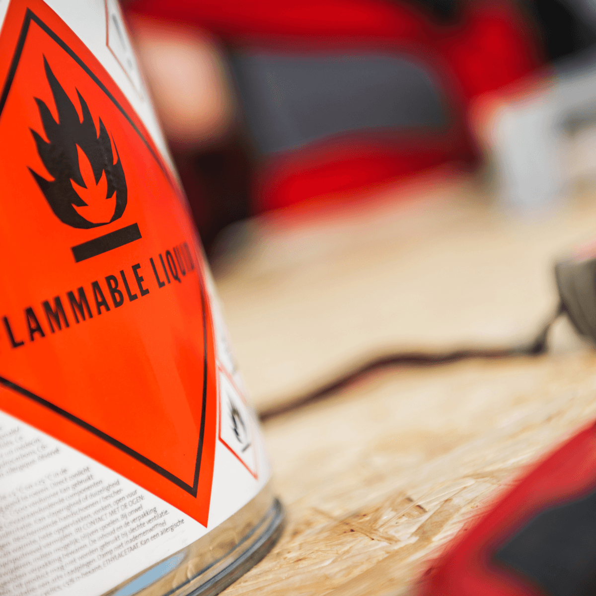 A close-up of a red diamond-shaped flammable liquid warning label on a glass container, set against a blurred industrial background.