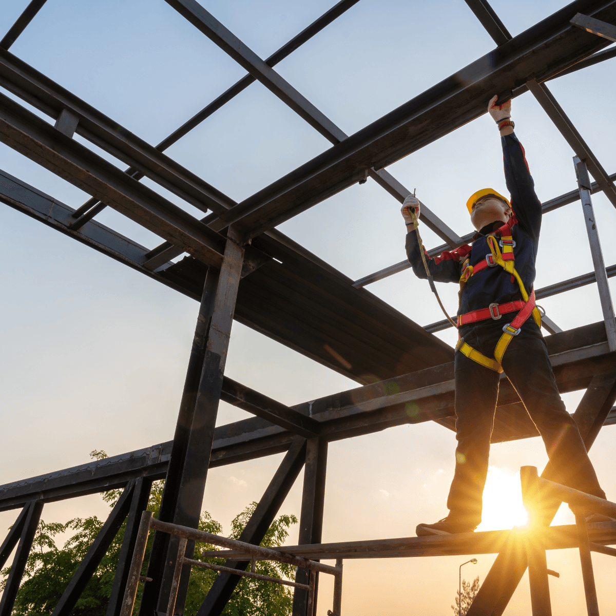A construction worker in a yellow hard hat and full safety harness working on a steel frame structure at sunset.