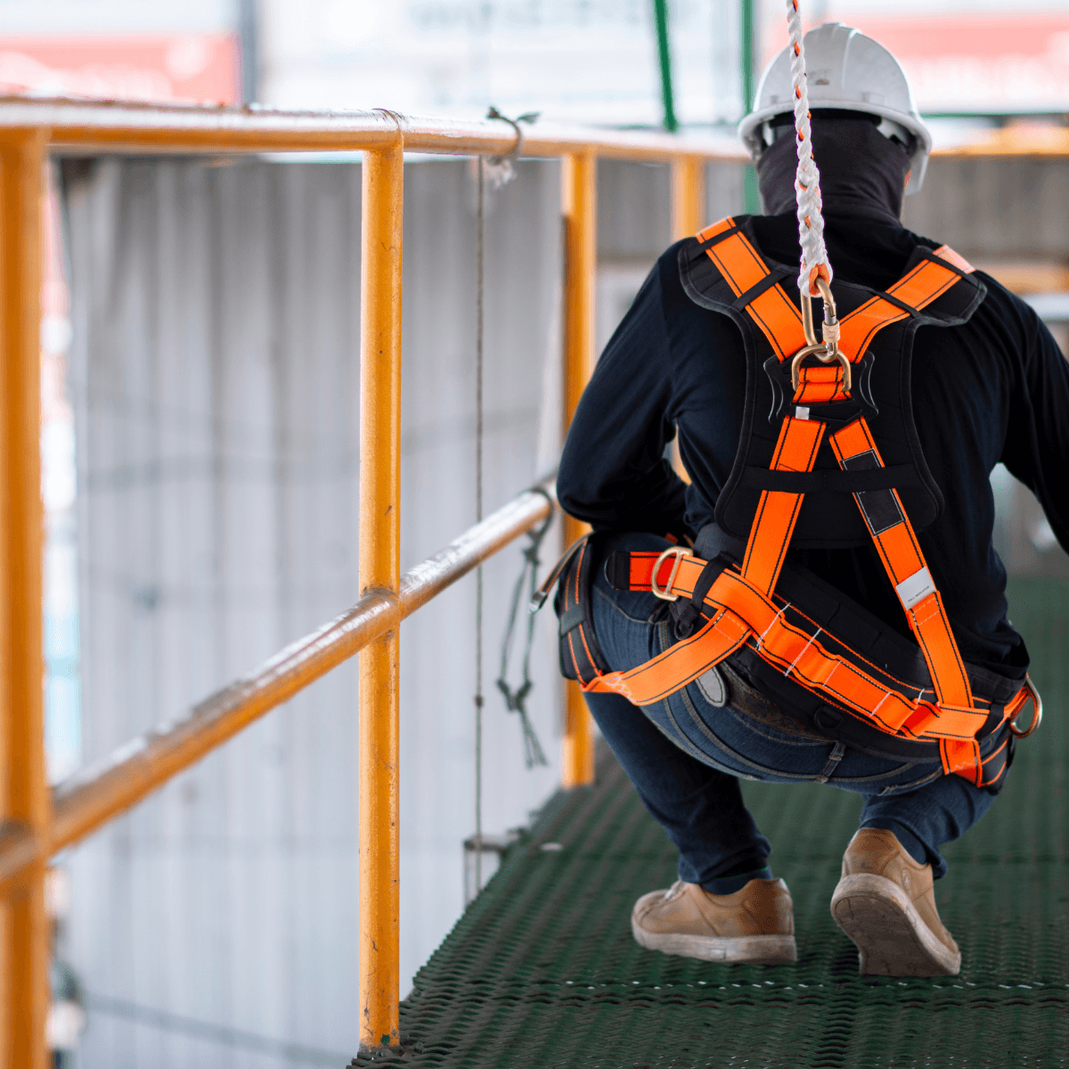 Worker crouched on an elevated platform, harness clipped to an overhead anchor line with a carabiner.