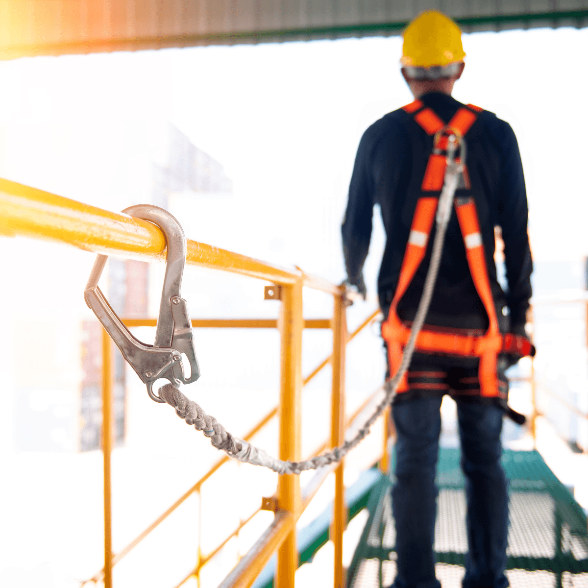 Close-up of a safety hook clipped to a yellow guardrail, with a worker in a harness standing in the background.
