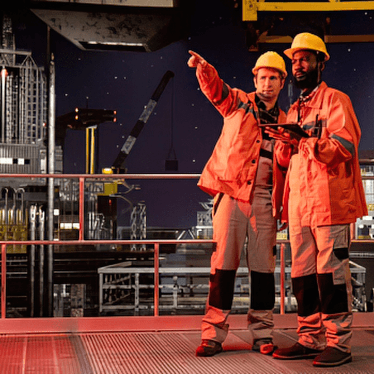 Two workers in orange coveralls and hard hats reviewing a tablet at an industrial site at night.
