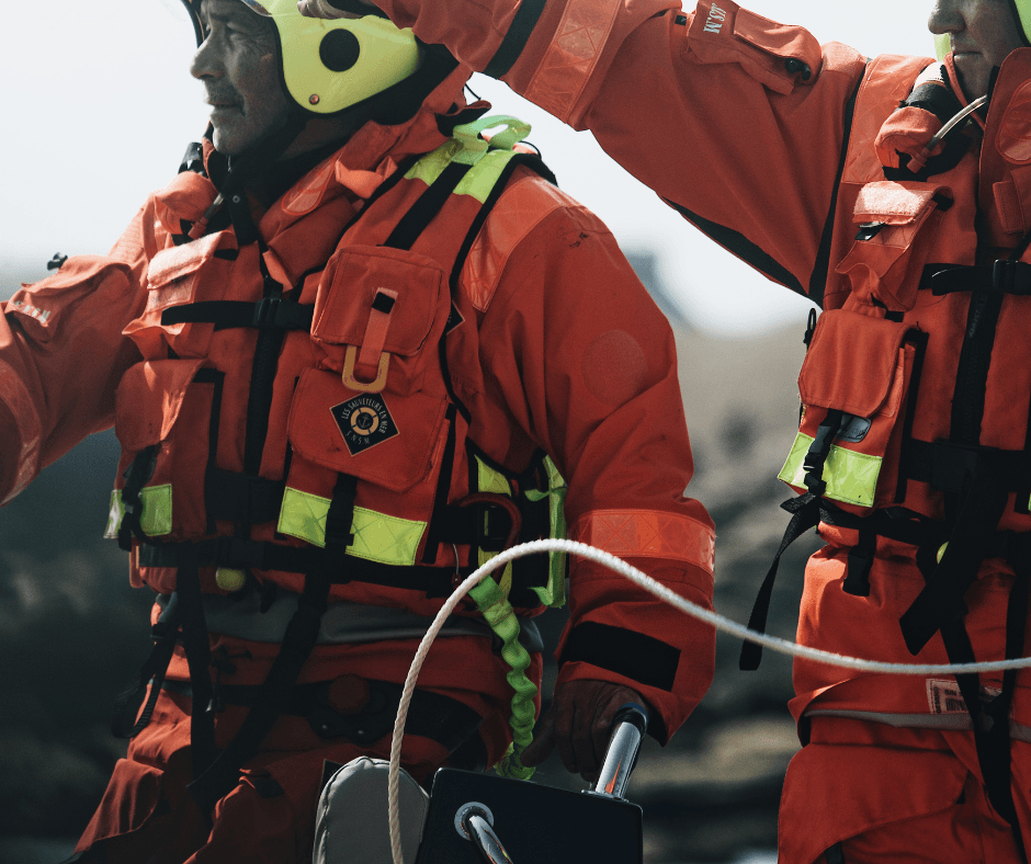 Rescue personnel in orange immersion suits with safety harnesses holding rope during marine rescue training